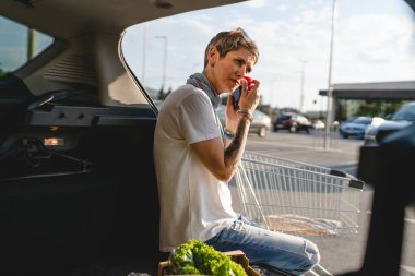 Yetişkin bir kadın arabasının bagajında oturan beyaz bir kadın süpermarketin otoparkında ya da markette vejetaryen ya da vejetaryen yiyeceklerle bir telefon görüşmesi yapıyor.