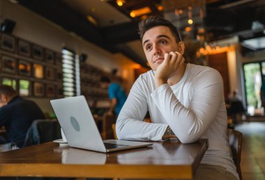 Kafe 'de dizüstü bilgisayarı olan genç, beyaz bir erkek. İnternette uzaktan kumandayla çalışıyor. Sorunları çözüyor.