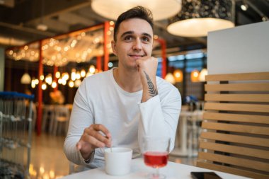 Genç, yetişkin, beyaz bir adam kafeteryada ya da restoranda tek başına oturuyor. Yan tarafa bakıyor, bir fincan kahve içiyor. Gerçek insanlar uzay gülüşünü kopyalıyor.