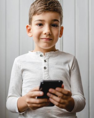 one boy caucasian child preschooler hold smartphone mobile phone at home play video games childhood and growing up technology addiction concept use smartphone app for online browsing or watch video