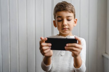 one boy caucasian child preschooler hold smartphone mobile phone at home play video games childhood and growing up technology addiction concept use smartphone app for online browsing or watch video