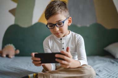 one boy caucasian child preschooler hold smartphone mobile phone at home play video games childhood and growing up technology addiction concept use smartphone app for online browsing or watch video
