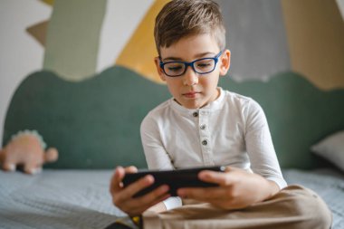 one boy caucasian child preschooler hold smartphone mobile phone at home play video games childhood and growing up technology addiction concept use smartphone app for online browsing or watch video