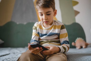 one boy caucasian child preschooler hold smartphone mobile phone at home play video games childhood and growing up technology addiction concept use smartphone app for online browsing or watch video