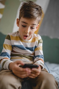 one boy caucasian child preschooler hold smartphone mobile phone at home play video games childhood and growing up technology addiction concept use smartphone app for online browsing or watch video