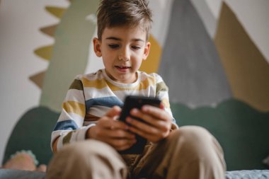 one boy caucasian child preschooler hold smartphone mobile phone at home play video games childhood and growing up technology addiction concept use smartphone app for online browsing or watch video