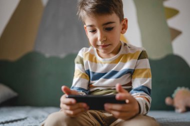 one boy caucasian child preschooler hold smartphone mobile phone at home play video games childhood and growing up technology addiction concept use smartphone app for online browsing or watch video