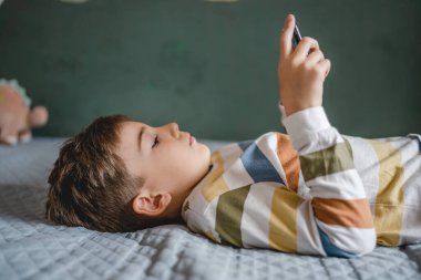 one boy caucasian child preschooler hold smartphone mobile phone at home play video games childhood and growing up technology addiction concept use smartphone app for online browsing or watch video