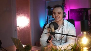 Bir beyaz erkek blogcu ya da vlogger yayın stüdyosunda video yayını yaparken el kol hareketi yapıyor mikrofon ve kulaklık kullanıyor.