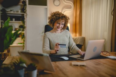 Modern beyaz bir kadın internetten alışveriş yapıyor. Dizüstü bilgisayar ve kredi kartı kullanıyor. Evinden mutlu bir gülümseme.