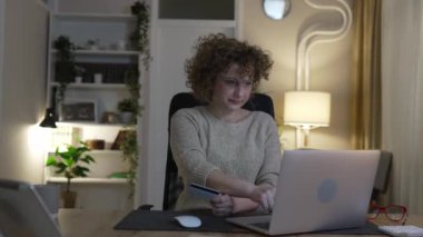 Modern beyaz bir kadın online alışveriş yaparken dizüstü bilgisayar ve kredi kartı kullanıyor. Evinden mutlu bir gülümseme. Geceleri masada kıvırcık saçlarıyla yalnız oturuyor.