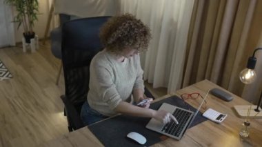 Modern beyaz bir kadın online alışveriş yaparken dizüstü bilgisayar ve kredi kartı kullanıyor. Evinden mutlu bir gülümseme. Geceleri masada kıvırcık saçlarıyla yalnız oturuyor.