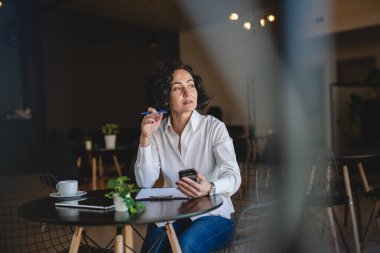 Bir kadın işkadını kafede ya da restoran yöneticisinin masasında oturuyor. İş yerinde akıllı cep telefonu kullanıyor. İnternette iş yapan olgun bir bayan girişimci.
