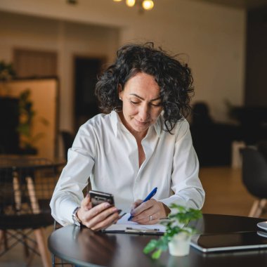 Bir kadın işkadını kafede ya da restoran yöneticisinin masasında oturuyor. İş yerinde akıllı cep telefonu kullanıyor. İnternette iş yapan olgun bir bayan girişimci.