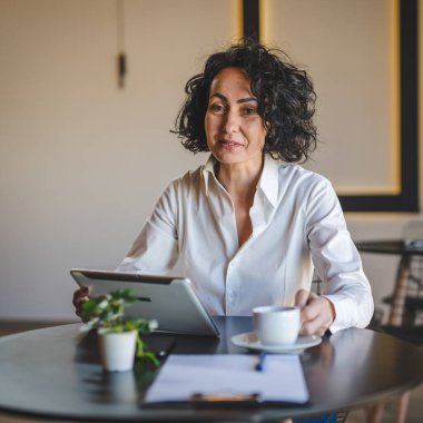 Bir kadın işkadını kafeteryada ya da restoran yöneticisinin masasında oturuyor. İş yerinde dijital tablet kullanıyor. İş yapan yetişkin bir kadın girişimci.