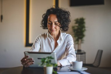 Bir kadın işkadını kafeteryada ya da restoran yöneticisinin masasında oturuyor. İş yerinde dijital tablet kullanıyor. İş yapan yetişkin bir kadın girişimci.