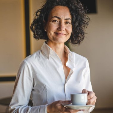 Bir kadın evdeki ya da işyerindeki pencerenin önünde duruyor. Girişimci kadın beyaz gömlek giyiyor. Kahve içiyor, gülümsüyor.