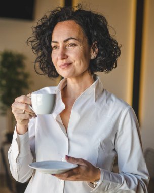 Bir kadın evdeki ya da işyerindeki pencerenin önünde duruyor. Girişimci kadın beyaz gömlek giyiyor. Kahve içiyor.