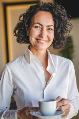 Bir kadın evdeki ya da işyerindeki pencerenin önünde duruyor. Girişimci kadın beyaz gömlek giyiyor. Kahve içiyor, gülümsüyor.