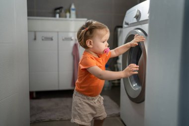 One girl small caucasian toddler child daughter standing at the washing machine in the toilet opening or closing the door examine and learn early development and growing up mischief concept copy space