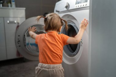One girl small caucasian toddler child daughter standing at the washing machine in the toilet opening or closing the door examine and learn early development and growing up mischief concept copy space