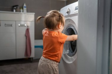 One girl small caucasian toddler child daughter standing at the washing machine in the toilet opening or closing the door examine and learn early development and growing up mischief concept copy space