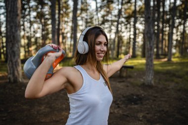 One woman adult caucasian female athlete using kettlebell girya weight during training in the forest park woods in summer day with headphones happy smile brunette health and fitness concept copy space