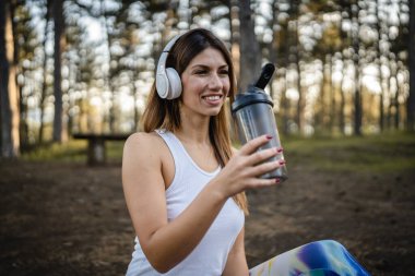 One woman happy caucasian female sitting in the forest or park in woods with supplement shaker and headphones during training in nature taking a brake water drink and hydration real people copy space