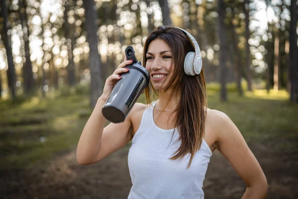 stock image One woman happy caucasian female standing in the forest or park in woods with supplement shaker and headphones during training in nature taking a brake water drink and hydration real people copy space