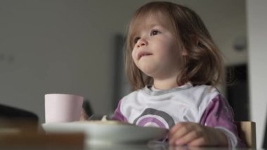 Küçük beyaz bir kız, yeni yürümeye başlamış bir kız çocuk. Evdeki masada yemek yiyor.
