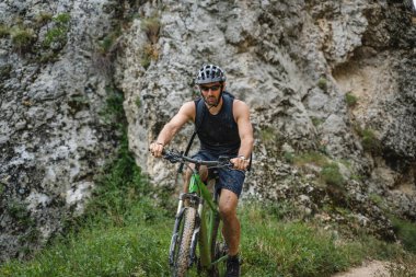 Kafkasyalı erkek bisikletçi, dağ geçidinde ya da tepenin eteklerinde elektronik bisiklet sürüyor. Güneşli bir günde koruyucu başlık ve gözlük takıyor.