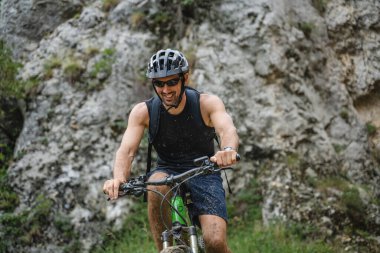 Kafkasyalı erkek bisikletçi, dağ geçidinde ya da tepenin eteklerinde elektronik bisiklet sürüyor. Güneşli bir günde koruyucu başlık ve gözlük takıyor.