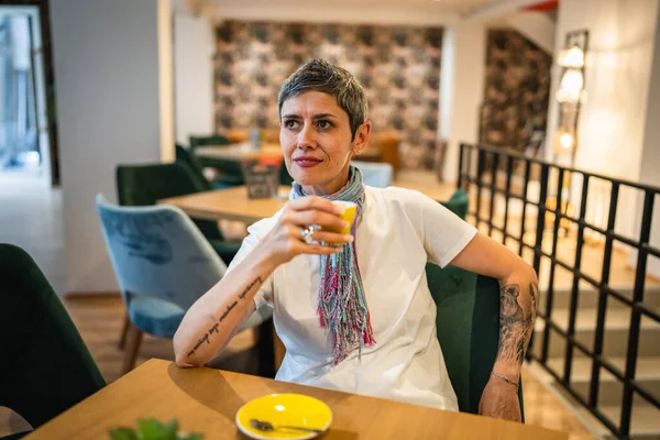 一位年长的高加索女人坐在咖啡店或餐馆里喝着一杯咖啡 真正的人模仿现代灰白短发人独自坐着的空间 — 图库照片