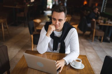 Kafe restoranındaki masada oturan beyaz tenli bir erkek bilgisayarla interneti tarıyor ya da uzaktan kumandalı bağımsız çalışıyor.