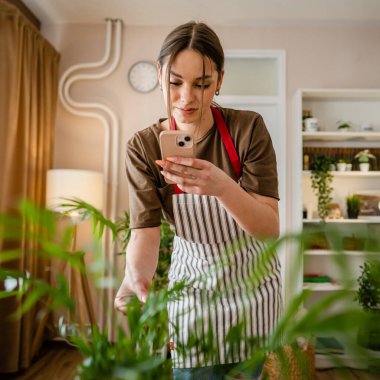 Bir kadın genç nesil z yetişkin beyaz kadın çiçekleriyle ilgileniyor akıllı telefonuyla saksı fotoğrafı çekiyor sosyal medyaya fotoğraf yolluyor ya da gerçek bir mesaj yolluyor.