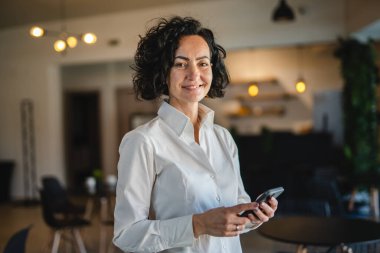 Yetişkin bir beyaz kadın mutlu bir gülümseme portresi kafede tek başına akıllı cep telefonunu tutuyor gerçek insanlar utangaç, beyaz gömlek giyiyor ve kıvırcık saçlı.