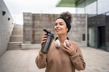 Kulaklıklı bir genç kadın eğitim için hazırlanıyor. Su içmek için destek şişesi tutuyor. Açık havada güzel beyaz kadın nesli.