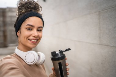 Kulaklıklı bir genç kadın eğitim için hazırlanıyor. Su içmek için destek şişesi tutuyor. Açık havada su içmek için güzel beyaz kadın selfie sağlıklı yaşam tarzı UGC Kullanıcı tarafından oluşturulmuş.