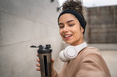 Kulaklıklı bir genç kadın eğitim için hazırlanıyor. Su içmek için destek şişesi tutuyor. Açık havada su içmek için güzel beyaz kadın selfie sağlıklı yaşam tarzı UGC Kullanıcı tarafından oluşturulmuş.