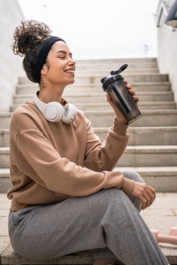 Kulaklıklı bir genç kadın eğitim için hazırlanıyor. Su içmek için destek şişesi tutuyor. Açık havada güzel beyaz kadın nesli.