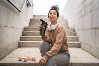 Kulaklıklı bir genç kadın eğitim için hazırlanıyor. Su içmek için destek şişesi tutuyor. Açık havada güzel beyaz kadın nesli.