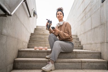 Kulaklık takan bir genç kadın destek şişesi ve akıllı telefon, spor, Kafkasyalı bir kadın Z jenerasyonu sağlıklı yaşam tarzı konseptini kopyalamak için fren yapıyor.