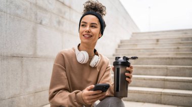 Kulaklık takan bir genç kadın destek şişesi ve akıllı telefon, spor, Kafkasyalı bir kadın Z jenerasyonu sağlıklı yaşam tarzı konseptini kopyalamak için fren yapıyor.