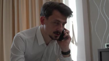 Yetişkin bir beyaz erkek otel odasından dizüstü bilgisayarla çalışıyor. İş gezisinde bıyıklı beyaz gömlek giyiyor. Uzaktan kumandalı iş adamı konsepti telefon konuşması yapıyor.