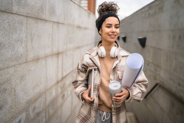 Genç bir kadın yetişkin kız öğrenci sınıfa gider dizüstü bilgisayarlı bir fincan kahve ve iş malzemesi taşır. Gerçek insan utangaç, mutlu, kulaklıklı, kendine güvenen bir gülümseme.