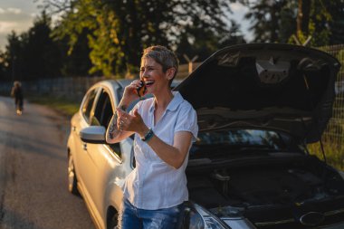 Olgun bir kadın akşamüstü yolda duruyor. Bozuk araba otomobili kaputu açık. Yol yardımı çekme konsepti için telefon ediyor.