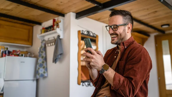 一名成年男子坐在沙发床上 身穿衬衫 戴着眼镜 坐在沙发上 用手机智能手机发送短信 或在网上浏览快乐的笑容 — 图库照片