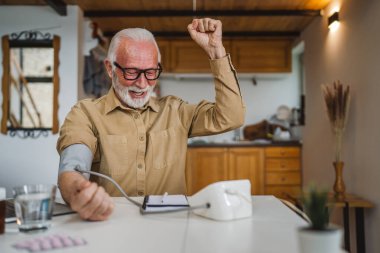 Mutlu, yaşlı, sakallı, sağlıklı beyaz bir erkek, kan basıncı cihazını kullanarak evde tek başına oturup sonuçları ölçüyor. Gerçek insanlar uzay sağlık hizmetleri konseptini kopyalıyor.