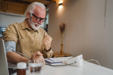 Mutlu, yaşlı, sakallı, sağlıklı beyaz bir erkek, kan basıncı cihazını kullanarak evde tek başına oturup sonuçları ölçüyor. Gerçek insanlar uzay sağlık hizmetleri konseptini kopyalıyor.
