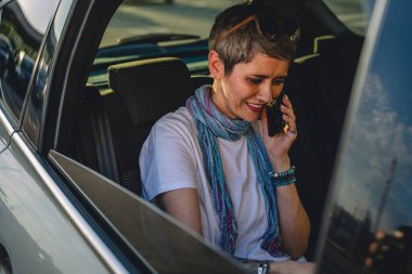 Yetişkin bir beyaz kadın, arabanın arka koltuğunda oturmuş dizüstü bilgisayarla çalışıyor. Yaz günü telefon görüşmesi yapıyor. Kısa gri saçlı, günlük fotokopi alanı takıyor.